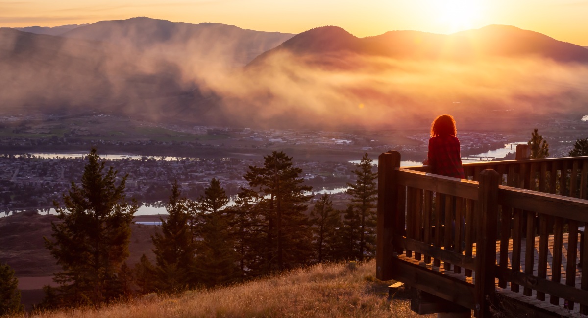 Girl standing on a lookout and looking at the city of Kamloops on a colourful summer with sunrise in the background.
