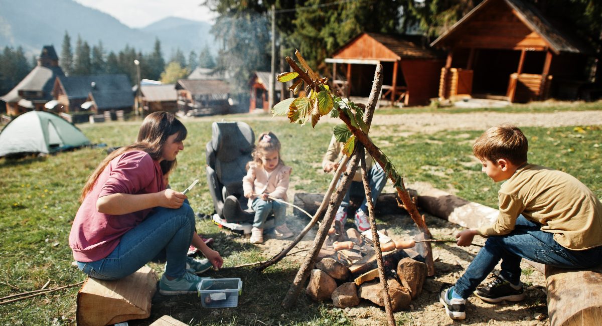 A family enjoying a bonfire in the mountains, with children roasting marshmallows and a backdrop of rustic wooden cabins surrounded by evergreen trees.